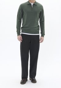 Pullover verde a maniche lunghe con colletto a mezza zip, abbinato a pantaloni neri a vestibilità ampia e scarpe marroni, presentato su uno sfondo neutro.