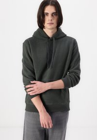 Hanorac din fleece verde închis, cu buzunar tip kangaroo, glugă cu șnur ajustabil, manșete cu nervuri și o textură moale, purtat cu pantaloni gri.