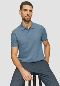 Herren Poloshirt mit kurzen Ärmeln aus weichem blauen Baumwollstoff. Verfügt über einen Kragen und eine Knopfleiste mit drei Knöpfen sowie ein kleines rotes Logo auf der Brust.