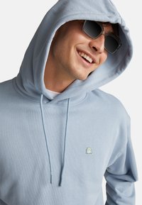 Felpa blu chiaro con cappuccio a coulisse, presenta un piccolo logo verde sul petto. Il tessuto appare morbido e liscio, con una vestibilità comoda.