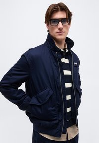 Jeune homme portant une veste bomber bleu marine avec une poche à rabat, un polo rayé, un sous-shirt beige, un jean foncé et de grandes lunettes teintées.