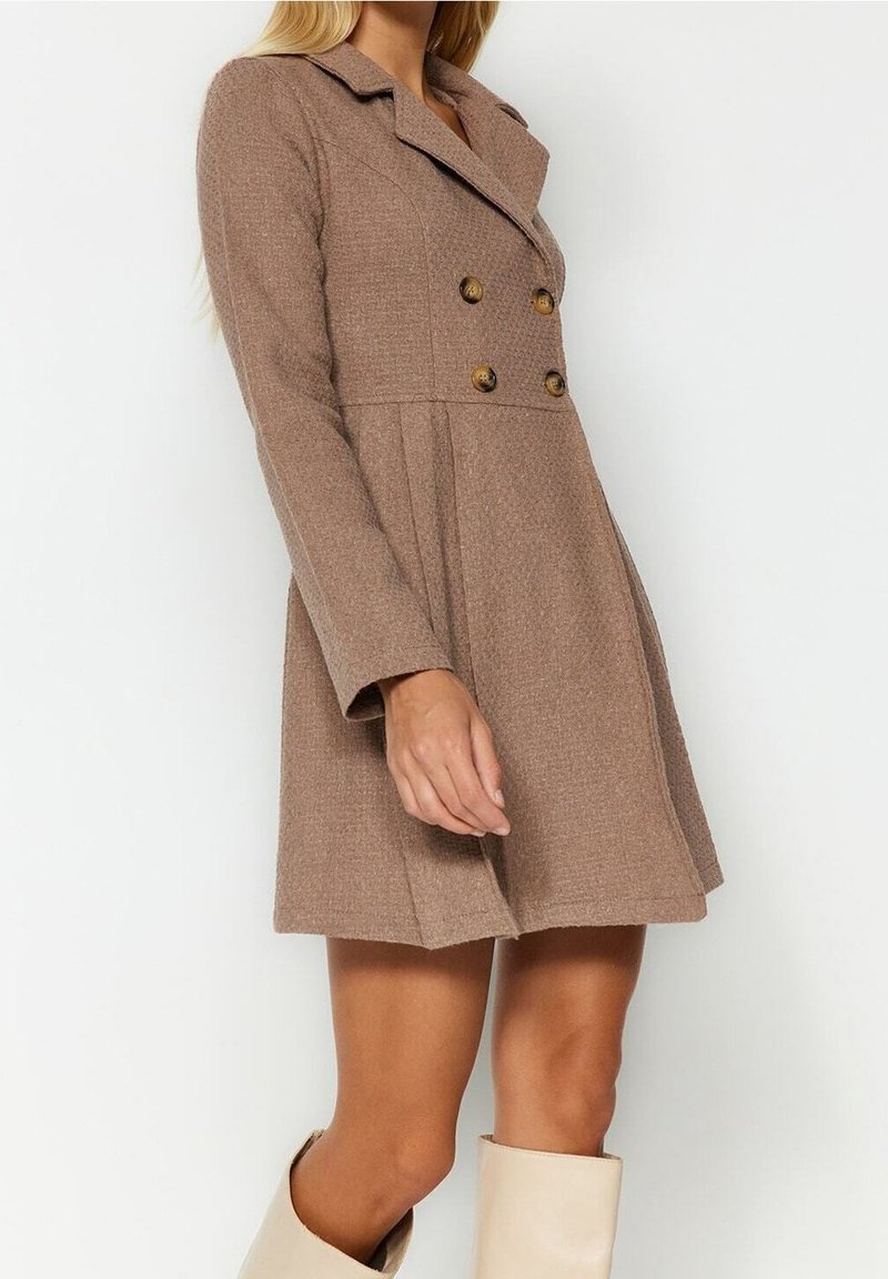 Femme portant un manteau en laine marron mi-longueur, à double boutonnage, manches longues, et des bottes beige montantes jusqu'aux genoux.