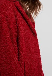 Červený kabát s texturovaným povrchem a chlupatou vnější stranou, s límcem ve stylu šály a otevřeným předním dílem. Tkanina působí měkce a teple.