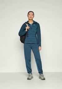 Prilagođena teal puffer jakna s zatvaračem, uparena s odgovarajućim tapered hlačama. Sive planinarske cipele i crni ruksak upotpunjuju odjevnu kombinaciju.