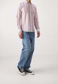 Homme portant une chemise à rayures roses et blanches avec boutons, un jean bleu et des baskets basses noires, debout devant un fond uni.