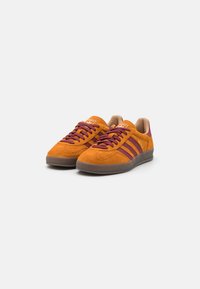 Baskets en daim orange avec des accents et des lacets bordeaux, présentant trois rayures bordeaux sur le côté, bout rond et semelle en caoutchouc.