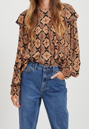 Femme portant un chemisier ample à motifs avec des épaules volantées et un jean bleu taille haute, main dans la poche.