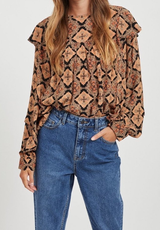 Femme portant un chemisier ample à motifs avec des épaules volantées et un jean bleu taille haute, main dans la poche.