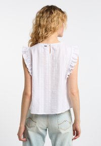 Blusa bianca a righe con maniche a volant e dettaglio a oblò sulla schiena, realizzata in tessuto leggero, abbinata a jeans di denim blu chiaro.