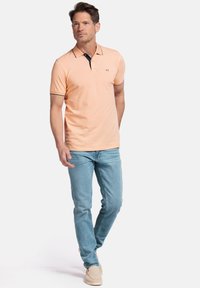 Polo en coton orange clair avec des accents de col marine, associé à un jean bleu clair et des chaussures beiges, se tenant devant un fond uni.
