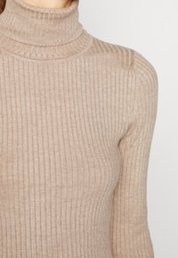 Beige värinen ribbineuleinen pooloneule, jossa on istuva malli ja teksturoitu kangas. Siinä on korkea kaulus ja pieniä ribbineuleisia yksityiskohtia kauttaaltaan.