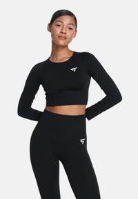 Crni sportski crop top s dugim rukavima i rebrastom teksturom, u kombinaciji s crnim tajicama visokog struka koje imaju bijeli logo.