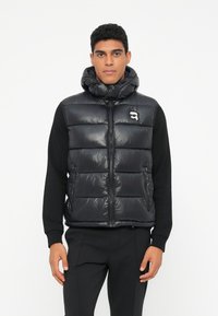 Melna pufer veste ar polsterētu dizainu, augstu apkakli, rāvējslēdzēju, divām sānu kabatām un mazu logo uz krūšu.