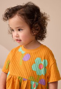 Vestido amarillo de manga corta con patrón acanalado texturizado, presenta grandes estampados de flores verdes y moradas, dobladillo en capas y cuello redondo.