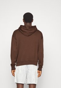 Felpa con cappuccio marrone, dal taglio rilassato, con polsini e orlo a coste. Indossata sopra pantaloni corti chiari e con fantasia.