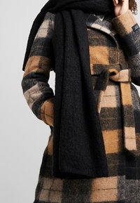 Manteau en mélange de laine à motif à carreaux marron, beige et noir. Associé à une longue écharpe noire texturée, maintenue par une ceinture.