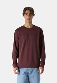 Felpa a maniche lunghe color bordeaux con scollo a girocollo, texture liscia e dettagli della cucitura frontale; abbinata a jeans di denim blu chiaro.