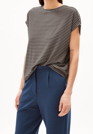 Personne portant un haut à manches courtes rayé gris et noir, rentré dans un pantalon bleu taille haute, sur fond blanc.