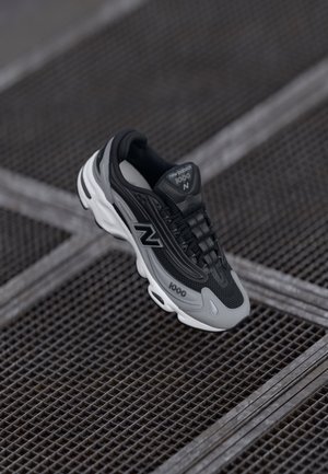 Czarny i szary sneaker New Balance 1000 z białą podeszwą spoczywający na chropowatej metalowej powierzchni z siatką.