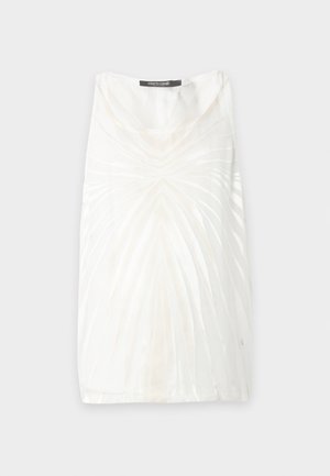 Man met een donkere huid die een wit topje met patroon zonder mouwen draagt, losse witte broek en zwart-witte loafers, staat voor een effen achtergrond.