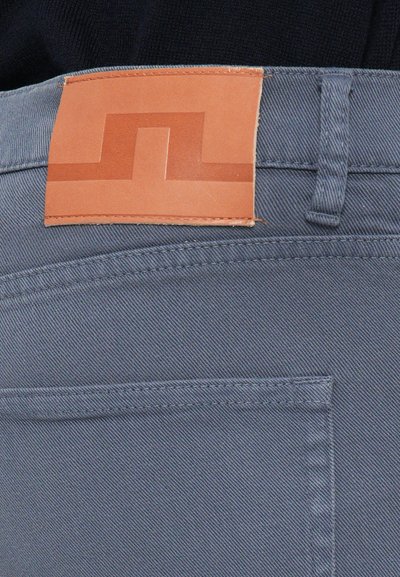Light grey denim jeans feature a tan leather waistband label with a geometric design. The fabric has a subtle texture and standard pocket detailing.
