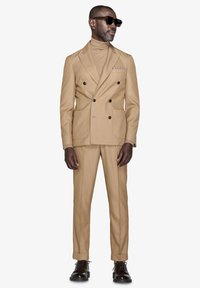 Beige dobbeltbrystet jakkesæt med skræddersyet pasform, matchende turtleneck og mørkebrune lædersko. Modellen bærer solbriller.