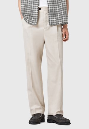 Pantalones beige ajustados con una textura suave, que cuentan con un corte relajado y bolsillos laterales. Combinados con una camisa gris a cuadros.