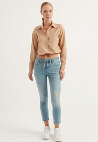 Camicia button-up marrone chiaro con maniche arrotolate, abbinata a jeans skinny strappati blu chiaro e sneaker bianche. Outfit semplice e casual.