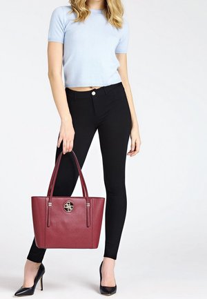 Femme en haut bleu clair à manches courtes et pantalon noir, tenant un grand sac à main bordeaux avec un logo doré, portant des talons hauts noirs.