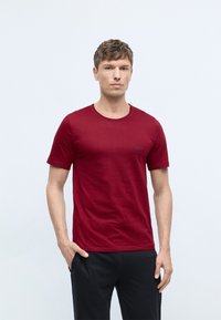 Burgundy cotton t-shirt with a round neckline, short sleeves, and a subtle logo on the chest. Worn with black pants, against a light background.