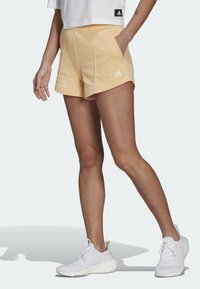 Persikofärgade träningsshorts med en mjuk textur, sidofickor och en kontrasterande vit Adidas-logotyp. Kombinerat med vita träningsskor.