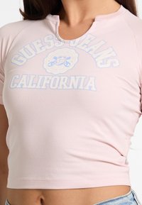 Hele roosa lühikesed varrukad särk ümarate kaelustega, millel on valge ja sinine "Guess Jeans California" logo, valmistatud pehmest ja venivast kangast.