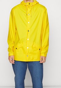 Person iført en lys gul regnjakke med trykknapper, snorehætte, klaplommer og blå denim jeans mod en ensfarvet baggrund.