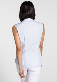 Blouse sans manches bleu clair avec une texture douce, coupe ajustée et un détail discret de couture au dos. Un pantalon blanc complète la tenue.