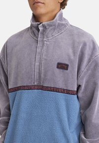 Mikina s fleecovým materiálom, ktorá má vrchnú časť z fialového kordu, spodnú časť z svetlo modrého fleece a vzorovaný tmavý pruh cez hrudník. Logo na vrchnej ľavej strane.
