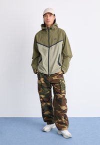 Letthvægtet jakke i olivengrøn og beige med lynlås og hætte, parret med camouflage cargo-bukser. Hvide sneakers fuldender det afslappede look.