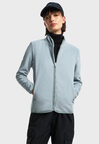 Svijetloplavi flis jakna s uspravnim ovratnikom i zatvaračem na munje. Sadrži bočne džepove i elastične manžete, nošena preko crnog rolaka.