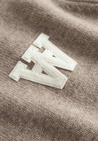 Beige strikket stof med en fremtrædende hvid broderet "A" og "N" logo med en tekstureret omrids. Nærbillede af syningens detaljer er synligt.