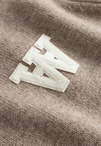 Tissu tricoté beige présentant un logo "A" et "N" brodé en blanc avec un contour texturé. Détail en gros plan des coutures visibles.