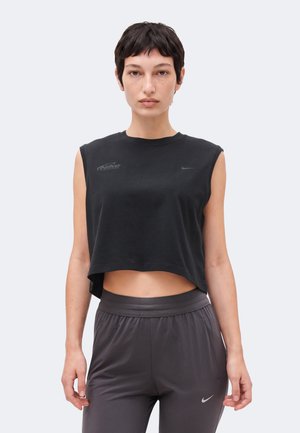 Jeune personne aux cheveux courts portant un haut court noir sans manches Nike et un pantalon de sport gris Nike, debout devant un fond uni.