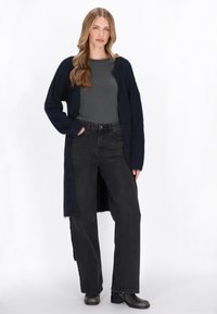 Hosszú, sötétkék kardigán, puha anyagból, amelyhez egy testhezálló szürke felsőt és fekete, magas derekú farmert visel. Fekete bokacsizmák is tartoznak hozzá.