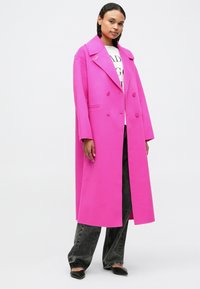 Sieviete, kas valkā spilgti rozā divrindas mēteli, melnus platās kājas džinsus, melnus norādītu kurpes un baltu apdrukātu topu uz vienkārša fona.