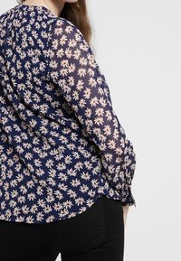 Mjõi blå topp med ett blommönster av vita och röda prästkragar, långa genomskinliga ärmar med rynkade manschetter, rundad nederkant och lätt tyg.