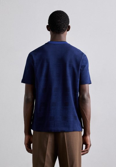 Mørkeblå t-skjorte med korte ermer og teksturert mønster. Crew neck-design, laget av myk stoff, vist fra baksiden. Brune bukser synlige.