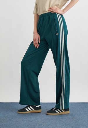 Personne portant un pantalon de survêtement Adidas teal avec des bandes blanches et des baskets Adidas noires avec des bandes blanches sur une moquette bleue.