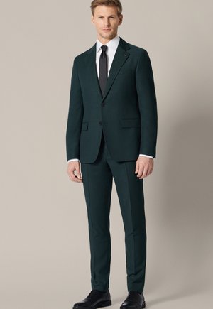 Mężczyzna ubrany w ciemnozielony garnitur typu slim fit, białą koszulę oraz czarny krawat, stojący na tle jednolitego jasnego tła.