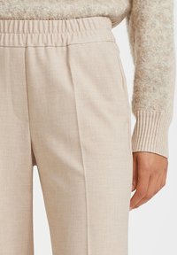 Beige housut korkea vyötärö, jossa on joustava vyötärönauha, sileä pinta ja sivulaskokset, esiteltynä kevyen neulotun neuleen kanssa.