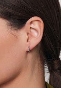 Złoty kolczyk w kształcie obręczy z fakturowaną powierzchnią, posiadający małe, fasetowane akcenty. Kolczyk noszony jest w przekutym uchu, widok z bliska.