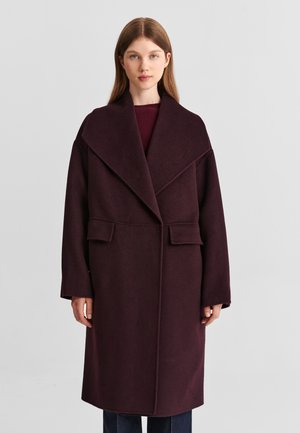 Jeune femme aux cheveux raides porte un long manteau en laine violet foncé avec de larges revers et des poches à rabat, debout devant un fond uni.