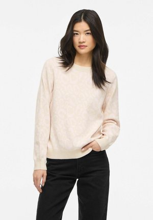 Jeune femme aux longs cheveux foncés portant un pull léger beige à motifs et un pantalon noir, debout, une main dans la poche.